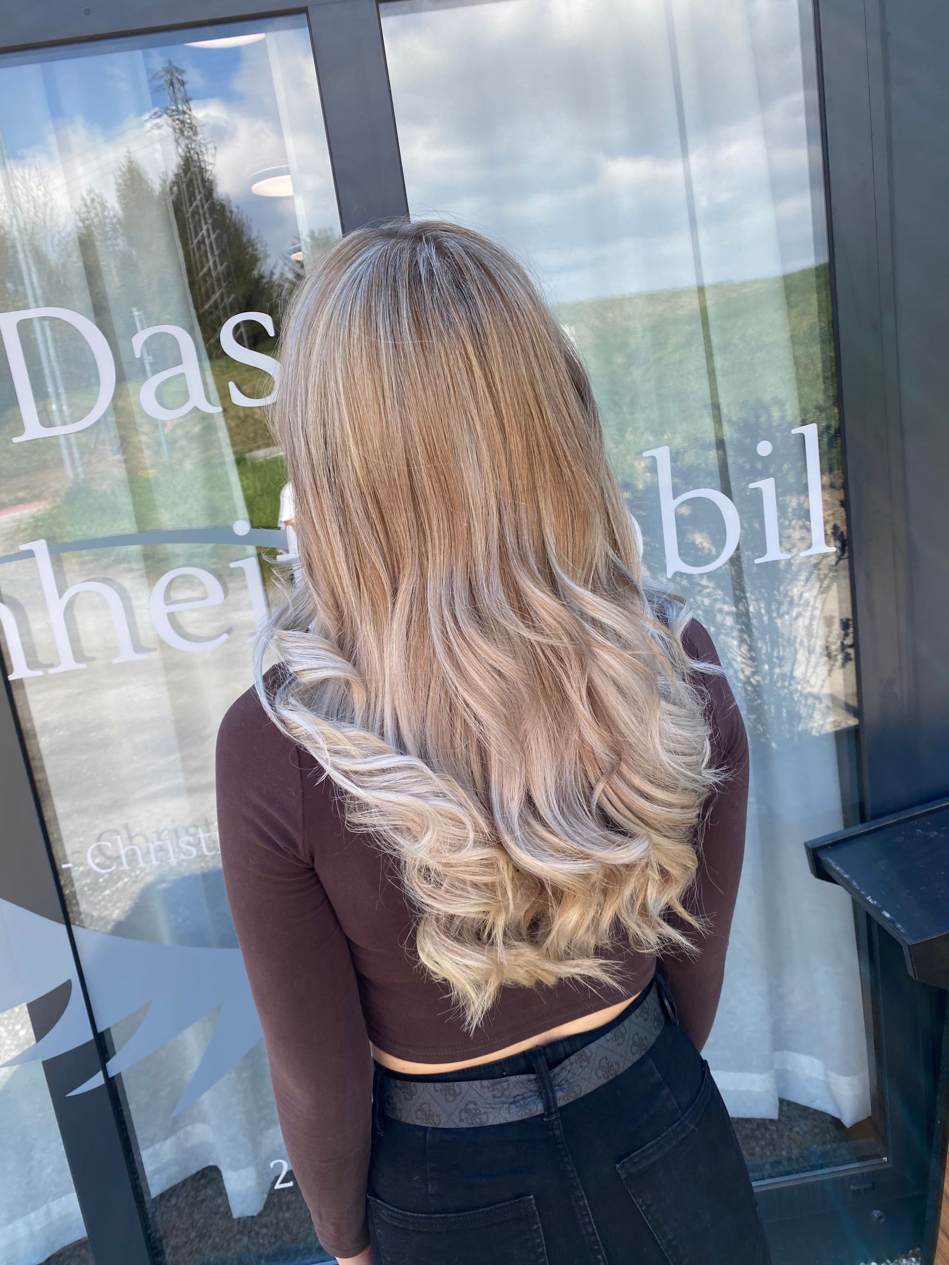 Eine Frau mit gewelltem, gekräuseltem blonden Haar steht vor einem Fenster. 