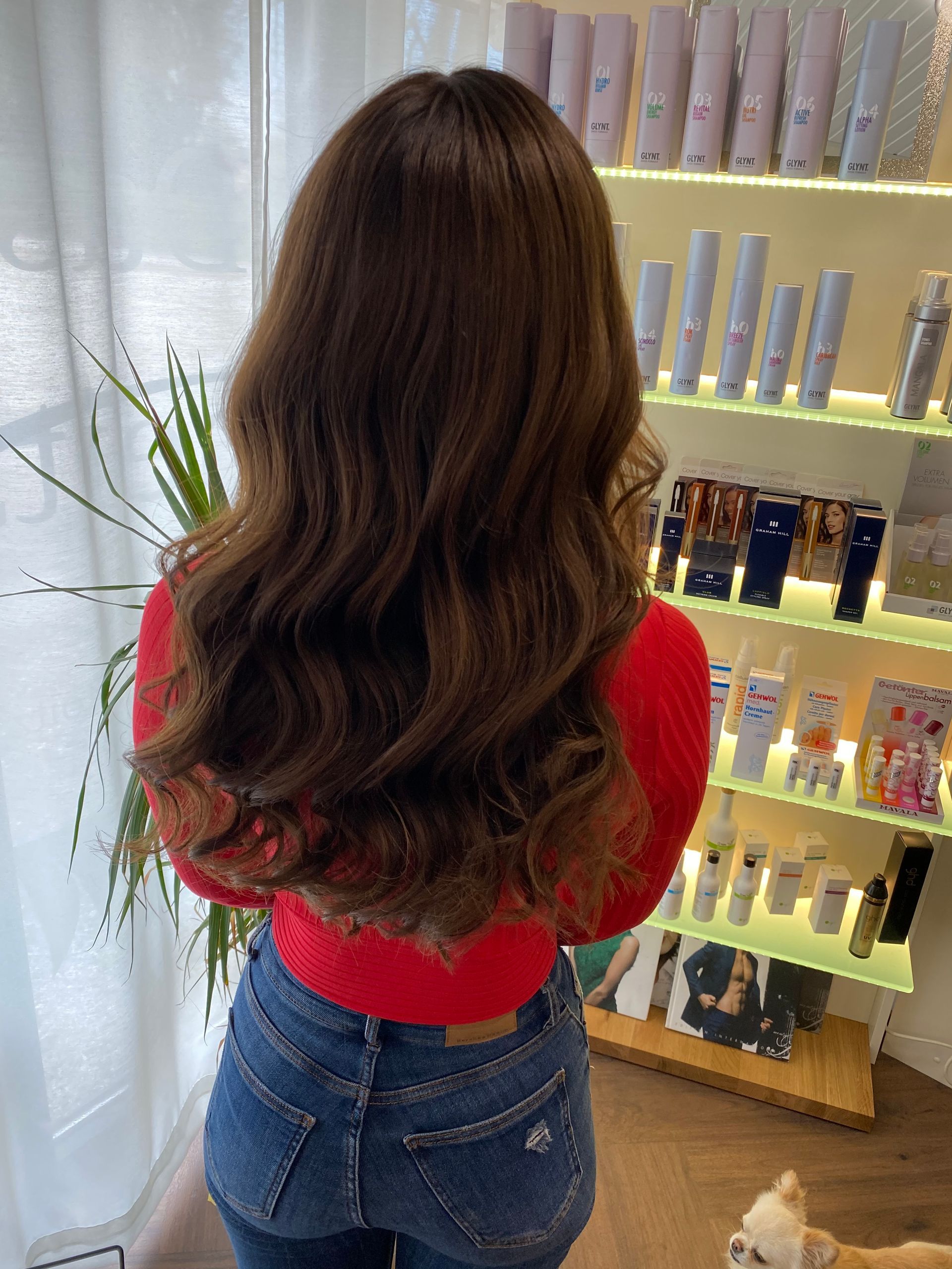 Person mit gewelltem braunem Haar steht in der Nähe eines Regals mit Schönheitsprodukten.