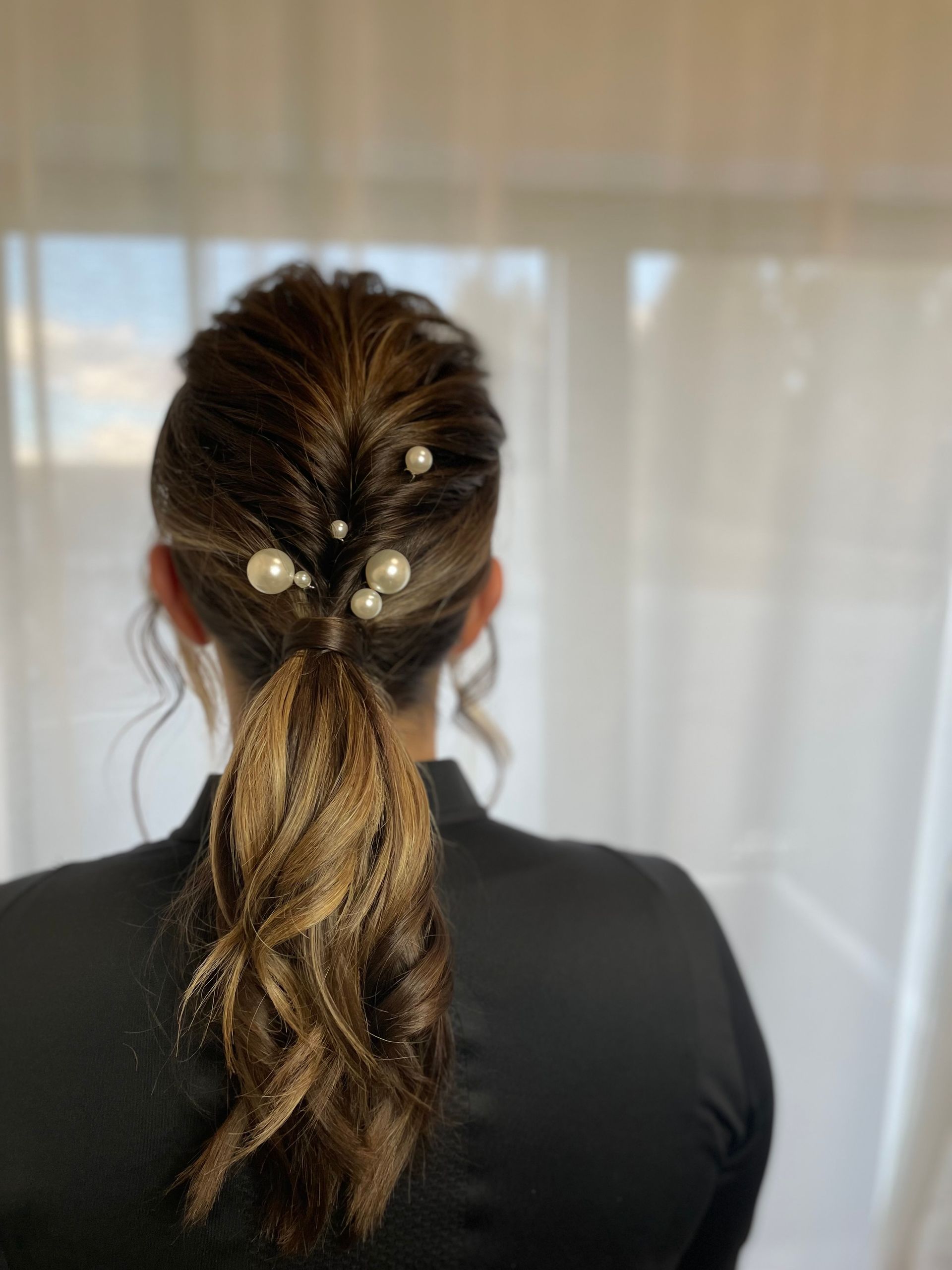 Die Haare der Frau sind zu einem Pferdeschwanz mit dekorativen Perlenspangen frisiert. Hellbraunes Haar mit dunklem Ansatz.