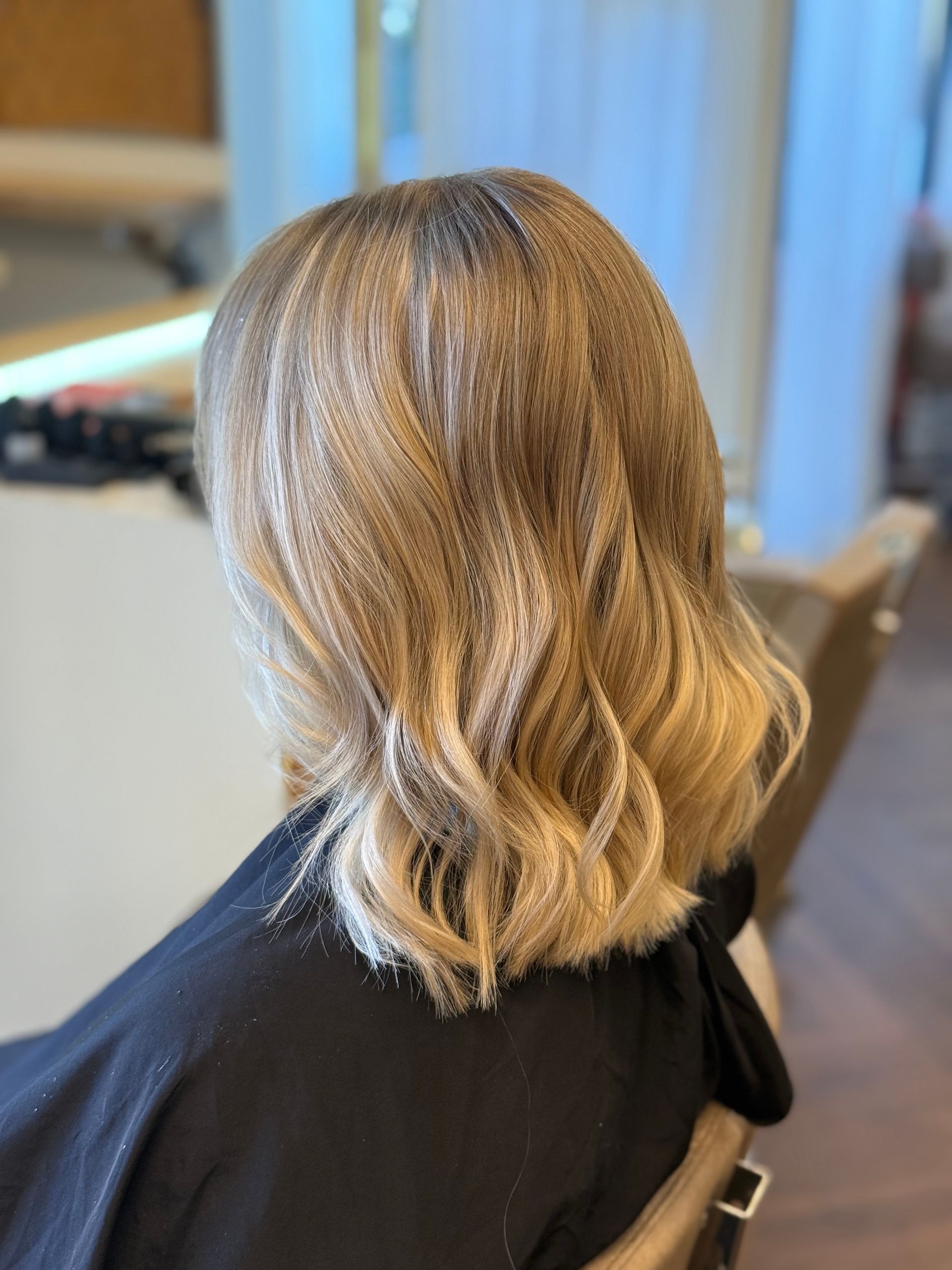Eine Person mit schultelangen Haaren sitzt in einem Friseurstuhl; die Haarwurzeln sind dunkel und gehen in ein Blond über.