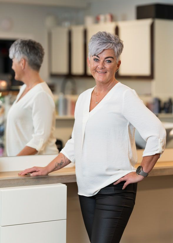 Frau mit grauem Haar lächelt und posiert mit einer Hand in der Hüfte in einem Salon. Weißes Oberteil, schwarze Hose.