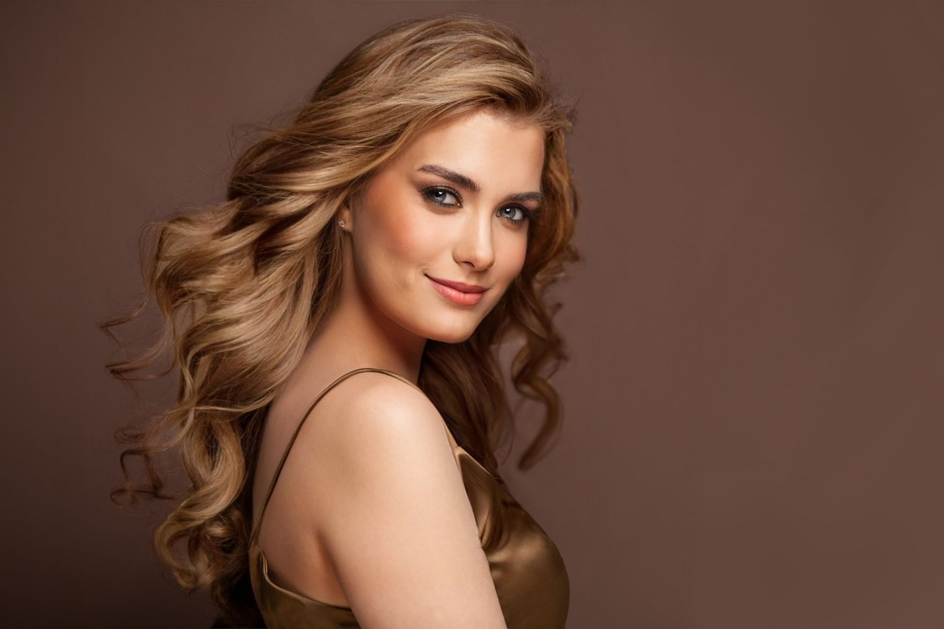 Frau mit welligem blonden Haar, lächelnd, in einem goldenen Kleid, vor braunem Hintergrund.