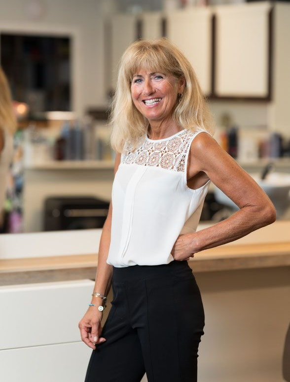 Frau mit blonden Haaren, die ein weißes Oberteil und eine schwarze Hose trägt, lächelt und posiert in einem Salon.