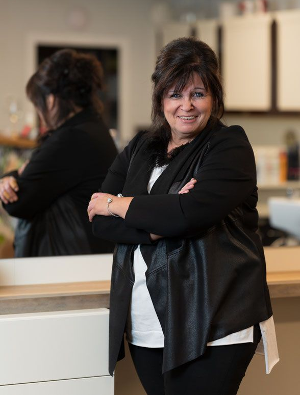 Frau in schwarzer Jacke und weißem Hemd, die Arme verschränkt, lächelt. Sie lehnt an einer Theke neben einem Spiegel.