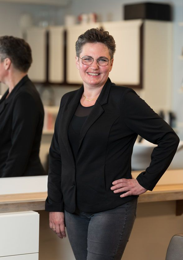 Frau mit kurzen Haaren, lächelnd, trägt Brille und schwarzen Blazer, die Hand in die Hüfte gestemmt