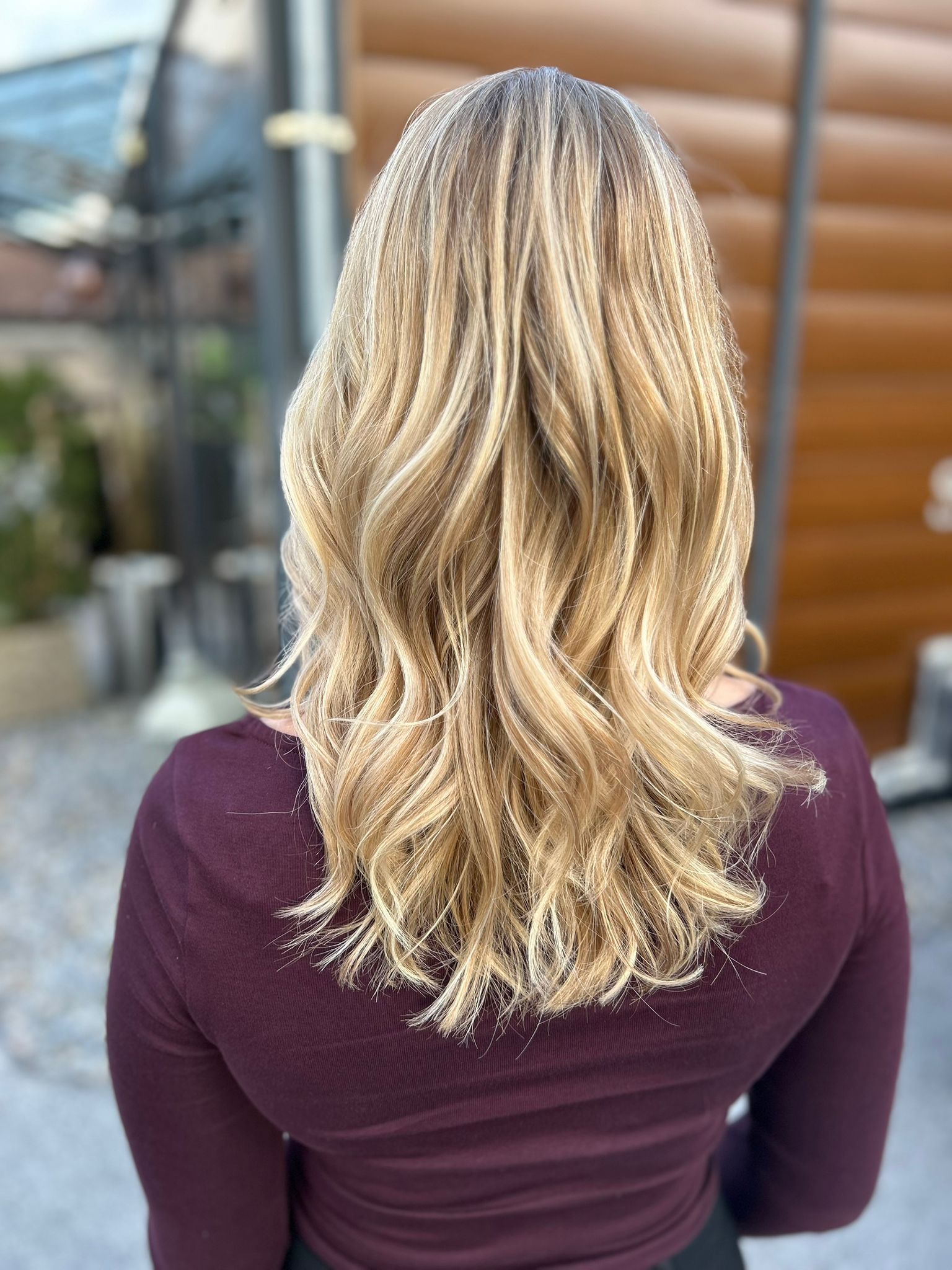 Frau mit langem, gelocketen blonden Haaren, die einen lilafarbenen Pullover trägt, im Freien auf einem Steinweg.
