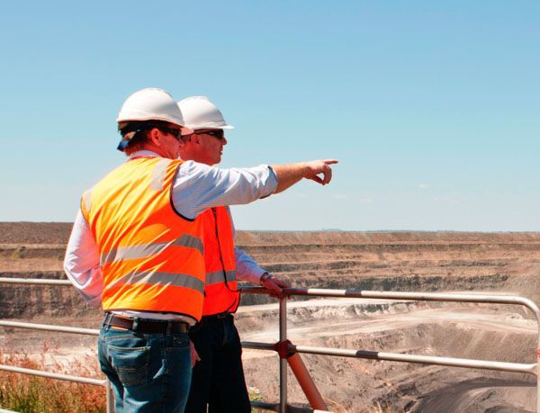Two men wearing hard hats and orange vests are pointing at something