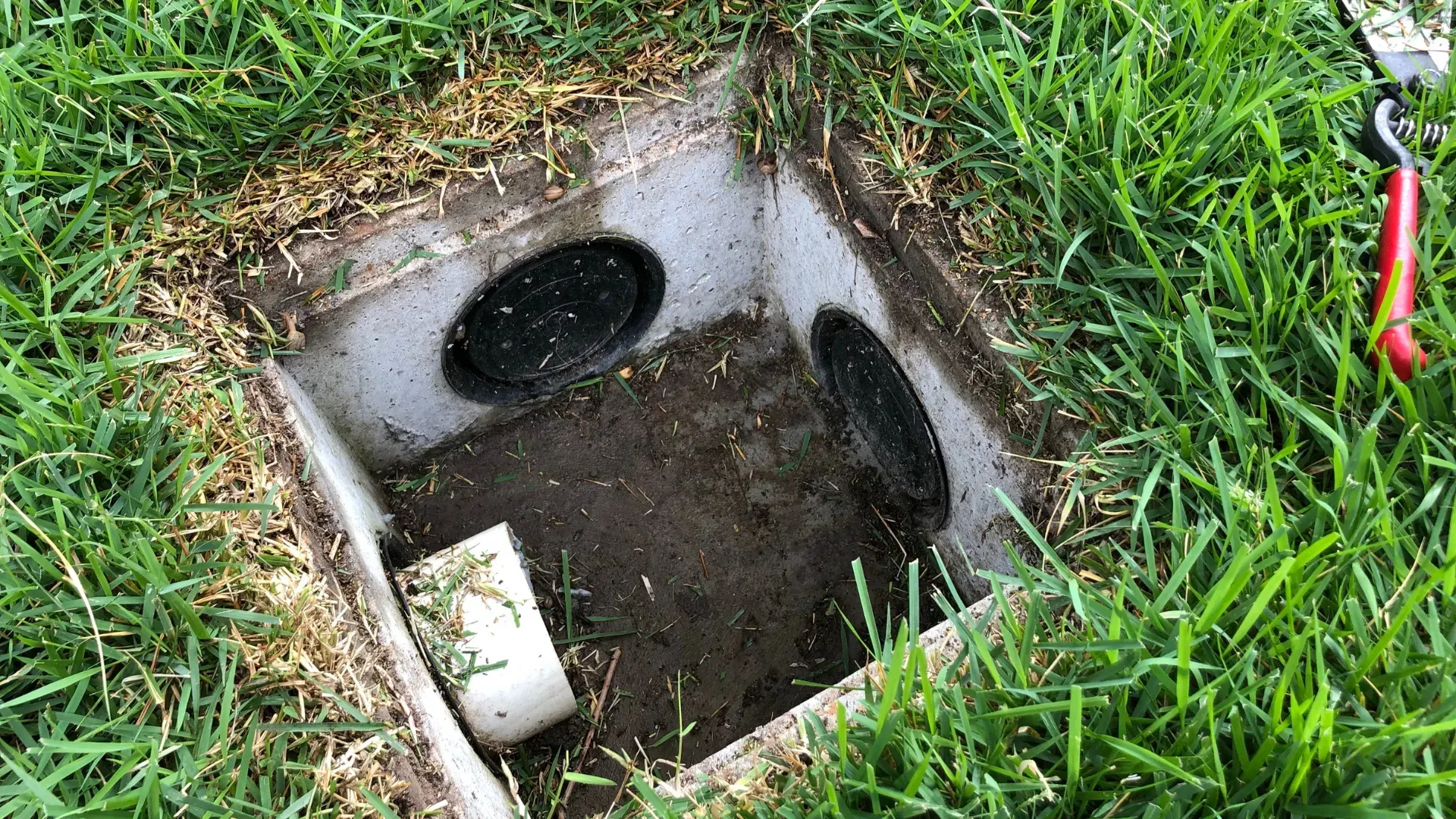 A square concrete irrigation valve box open in green grass, showing black pipes and a white PVC pipe.