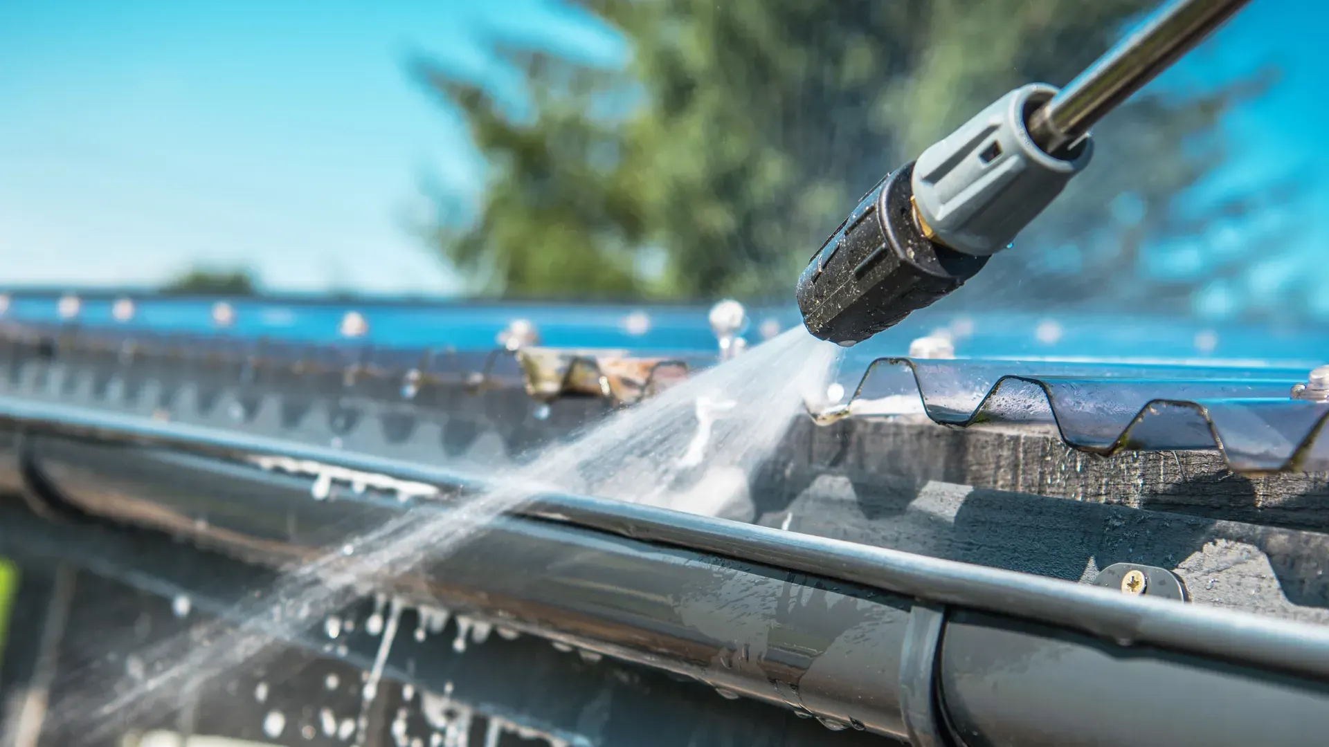 A pressure washer sprays water on a dark gutter, removing debris under a blue sky.