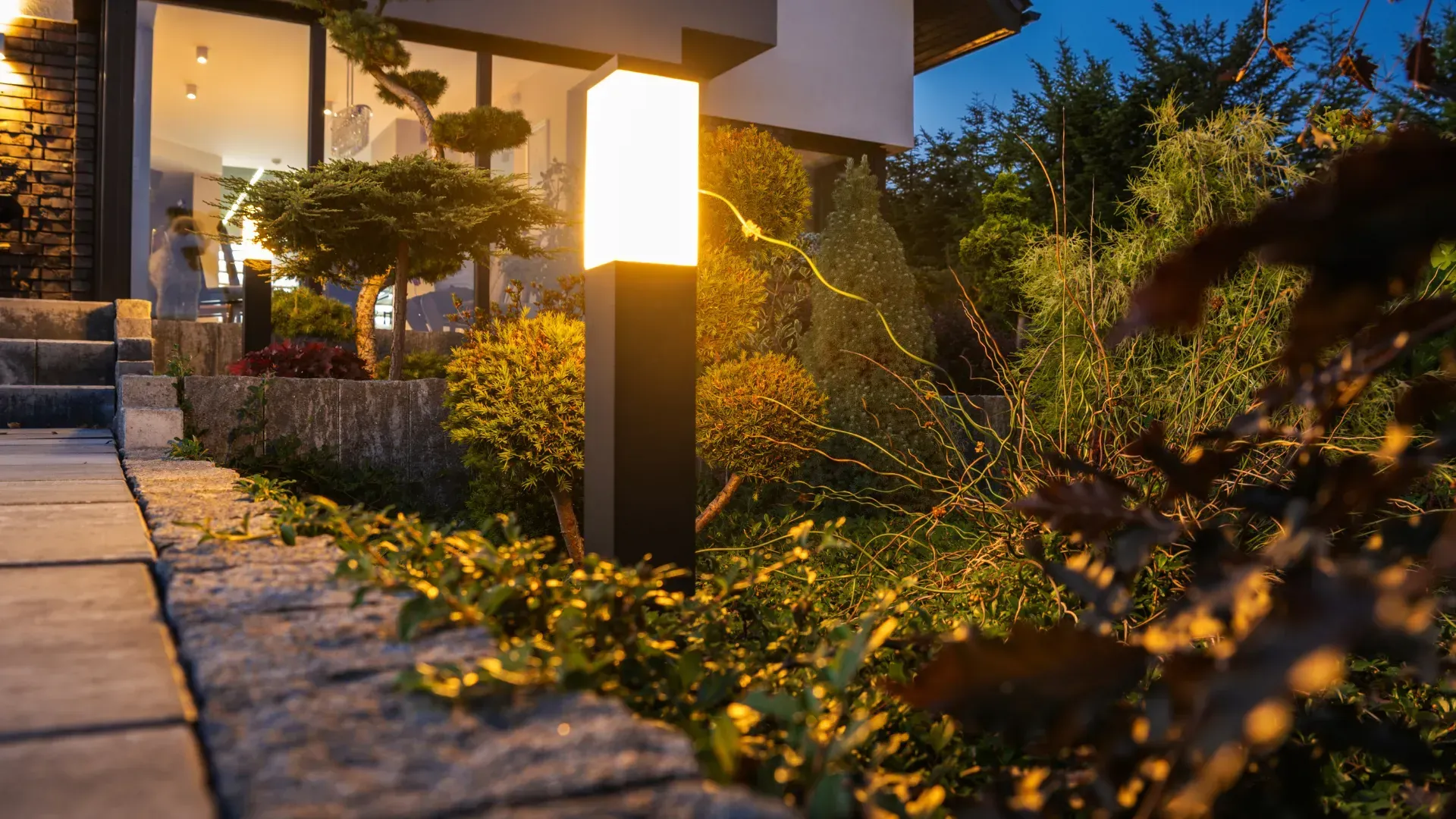 Lit outdoor lamp in a garden with landscaping and a building in the background at dusk.