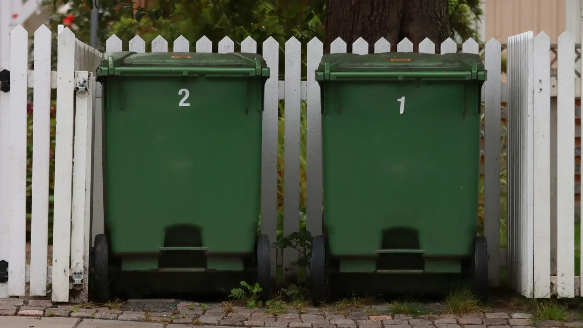 Two green trash bins with numbers 1 and 2, set between a white picket fence.