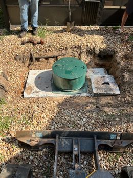 Workers near a septic tank, which is a green lid on a concrete pad.  A small excavator is in the foreground.