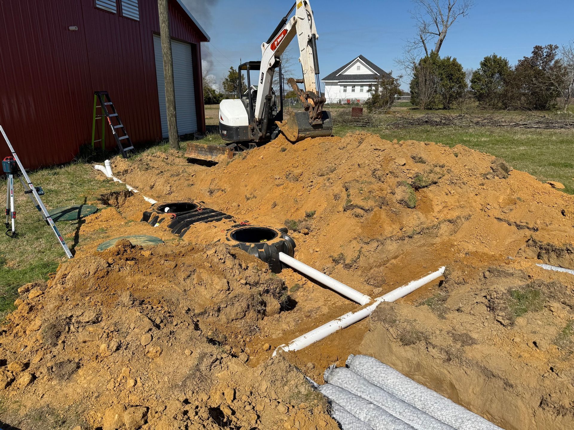 Construction site: Excavator near a red barn and a white house, with installed septic system components and buried pipes.