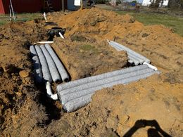Construction site, septic tank installation; pipes, excavated soil, black tank, red building in the background.