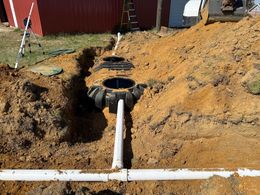 Construction site, septic tank installation; pipes, excavated soil, black tank, red building in the background.