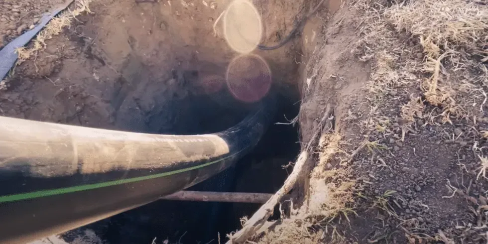 A black pipe extending into a dark hole dug in the ground. Sunlight glares.
