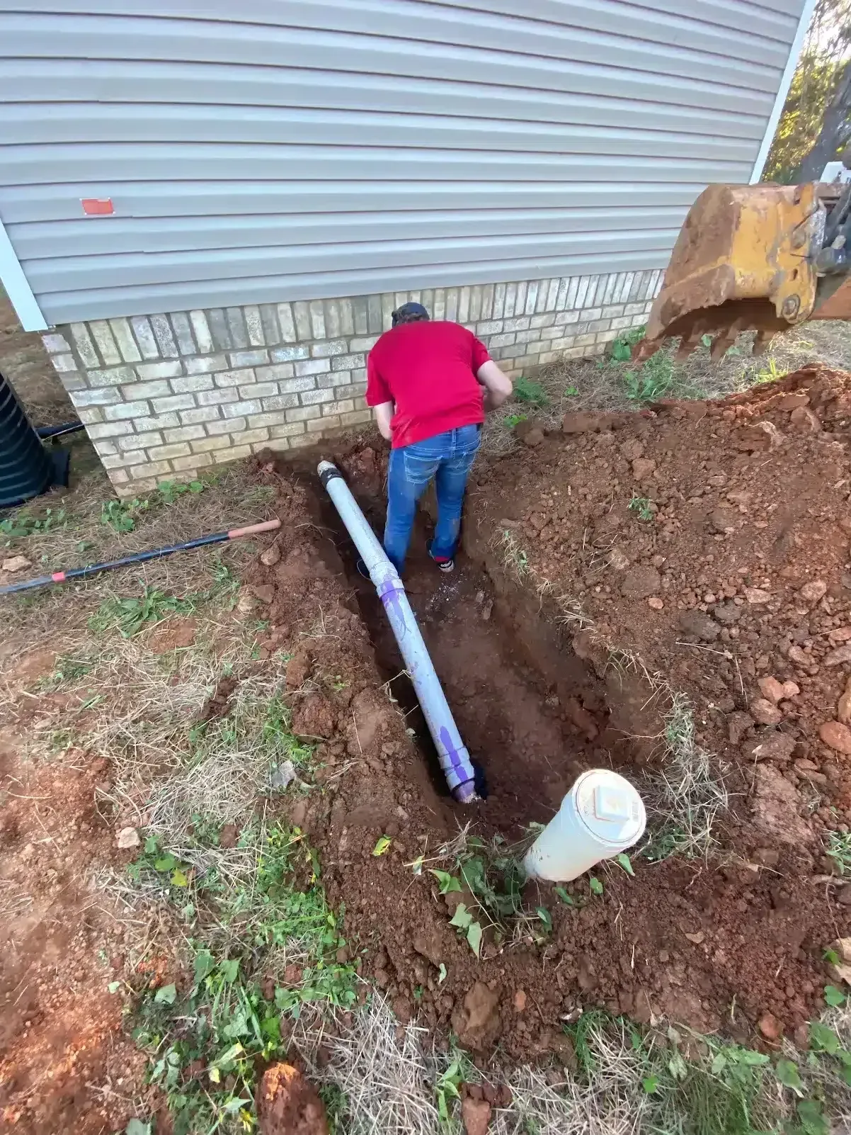 A person in a red shirt works on a pipe in a trench near a house; soil, grass, and machinery are nearby.