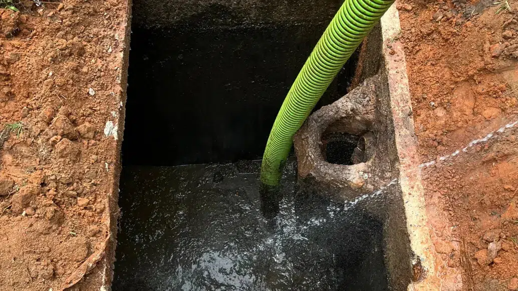 Green hose spraying water into a dark, brick-lined pit in an outdoor setting.