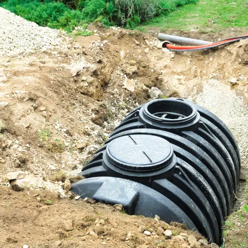 Black septic tank partially buried in a dirt trench; two access lids visible; setting is outdoors.