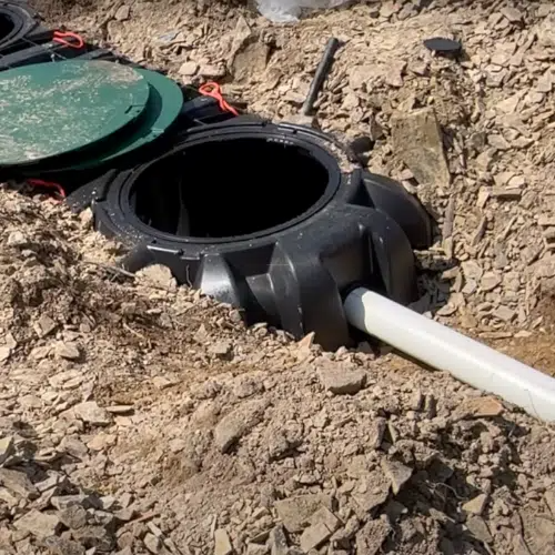 Black septic tank with open lid, buried in dirt, with white pipe connected.