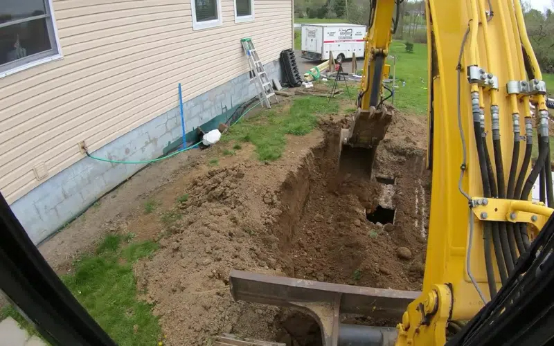 Yellow excavator digging trench next to a house with beige siding and green grass.