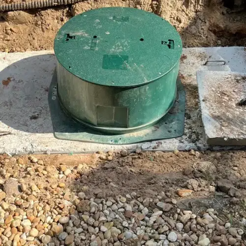 Green circular septic tank lid set on a concrete base next to a rectangular access lid; outdoor setting.