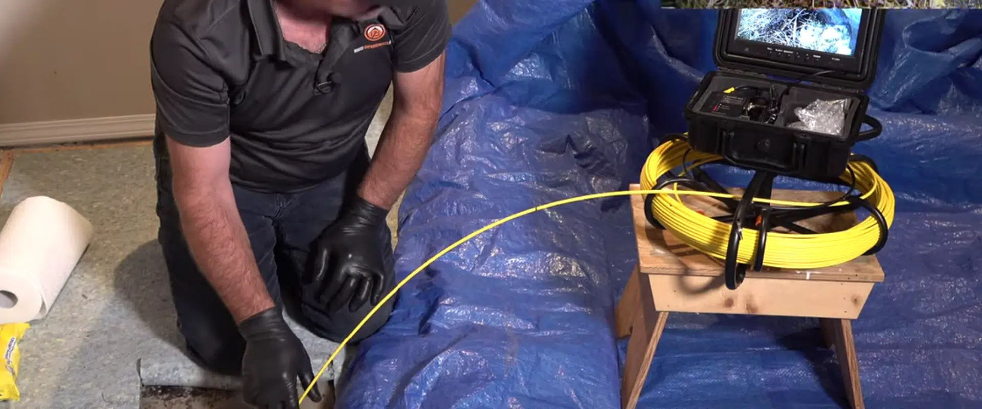 Man in gloves inspects plumbing with a camera on a blue tarp, next to a yellow cable.