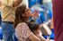 Una mujer, sentada entre sillas azules, sostiene a un niño en brazos, mira hacia arriba y aplaude.