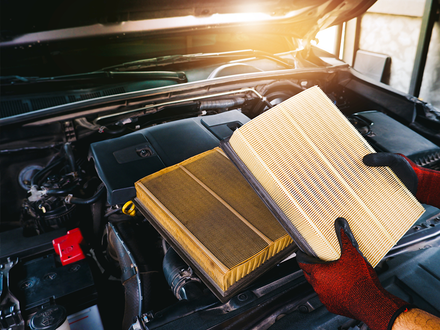 A person is holding two air filters in front of a car engine.