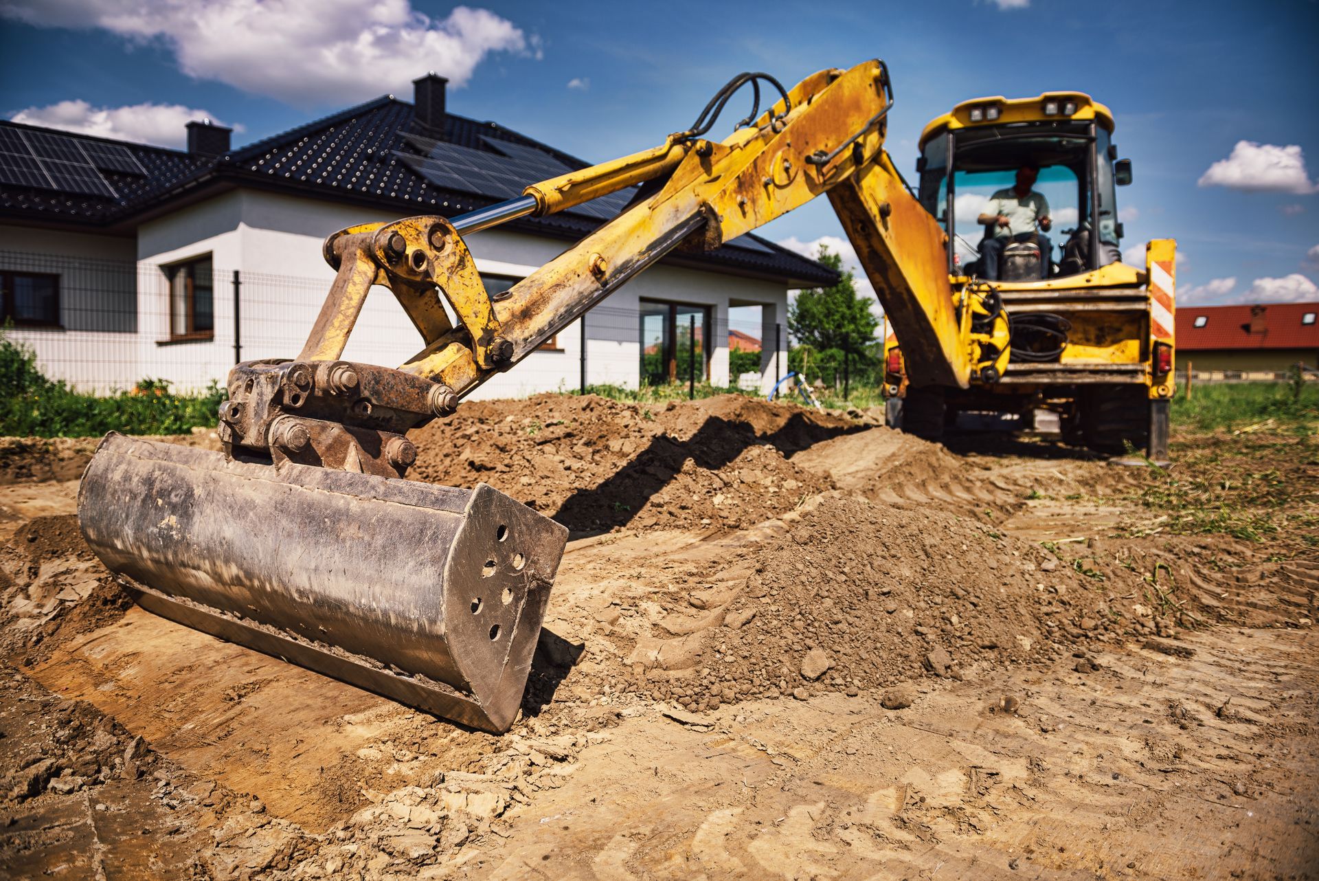 Yellow excavator digging a trench at a construction site near a white house on a sunny day.