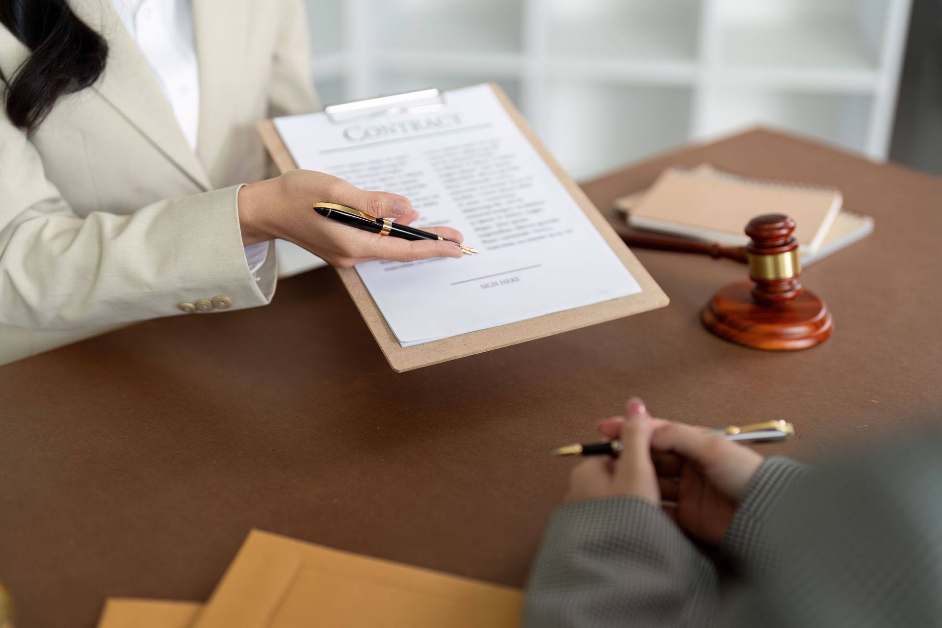 Person in a blazer presenting a contract with a pen to another person. A gavel and books are on the table.
