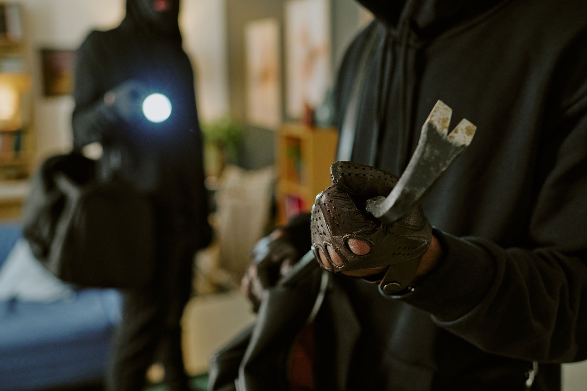 Two people in black hoodies inside a house, one holding a crowbar, the other a flashlight and bag.