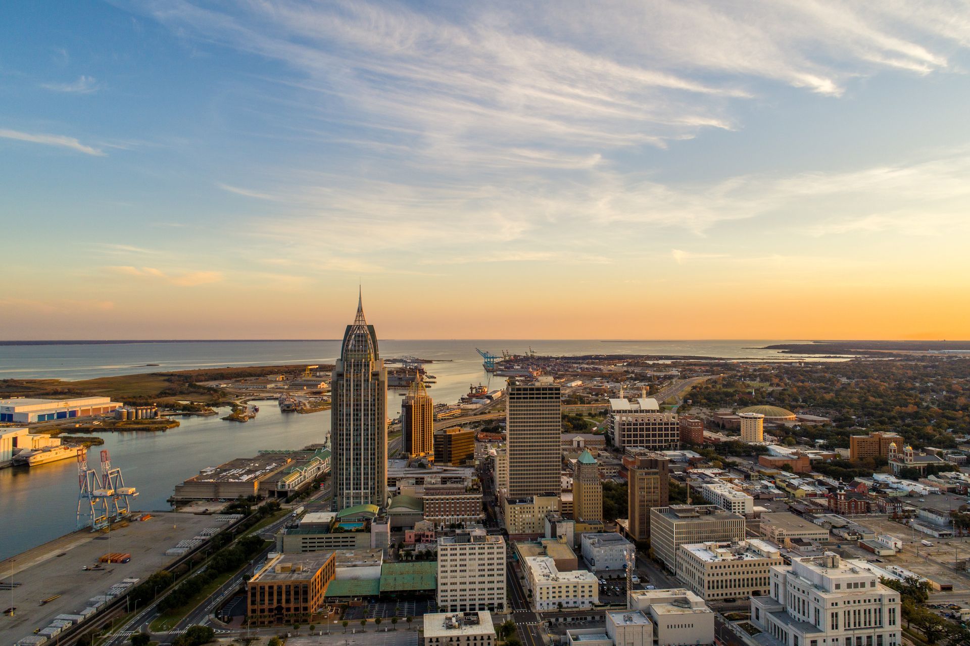 Aerial view of Mobile, Alabama, at sunset; tall buildings, waterfront, and orange-tinted sky.