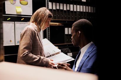 Woman and man reviewing documents in an office. The woman wears a blazer, man wears a suit. Shelves and binders visible.