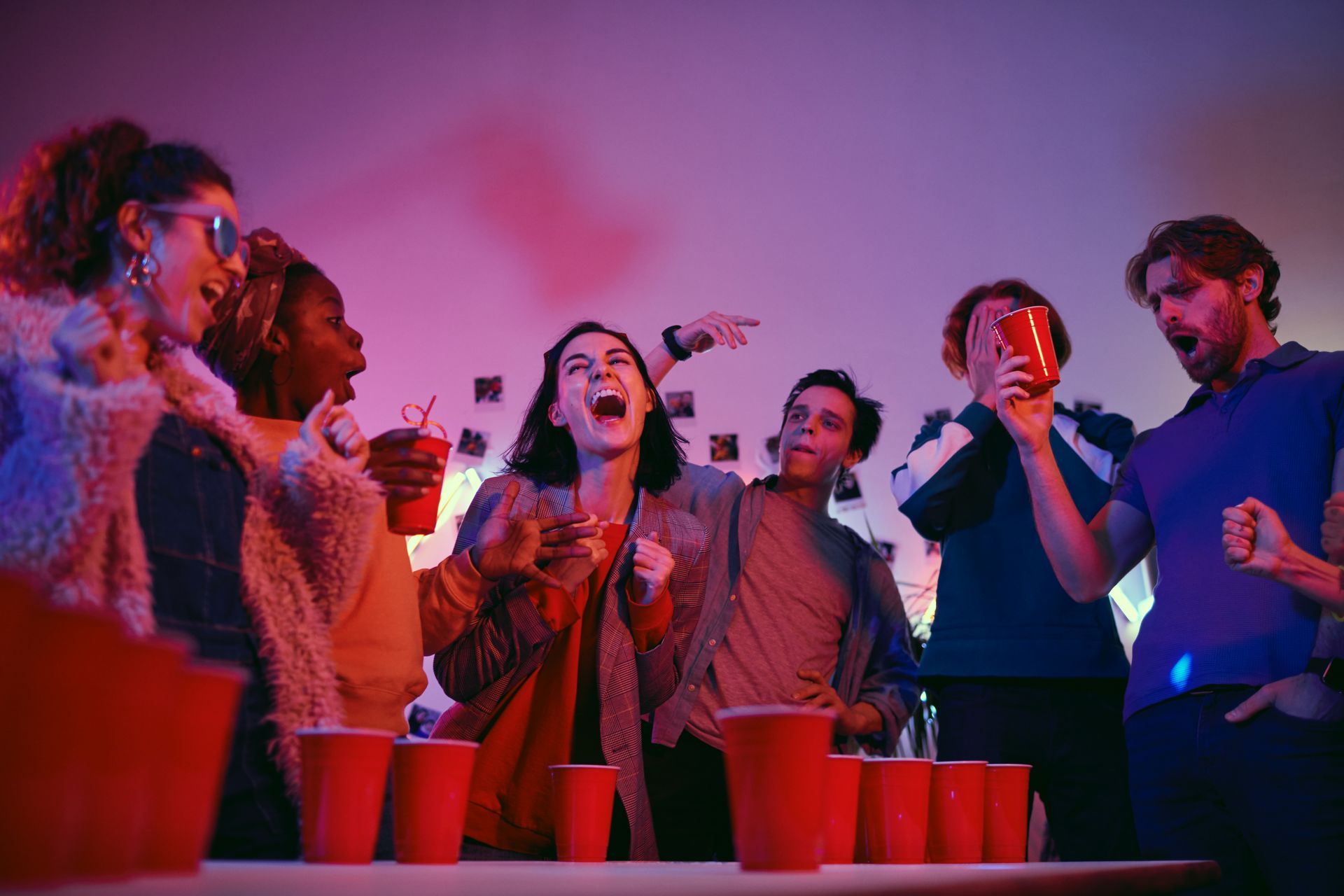 Friends celebrating, playing a game with red cups. Purple and pink lighting illuminates their excited expressions.