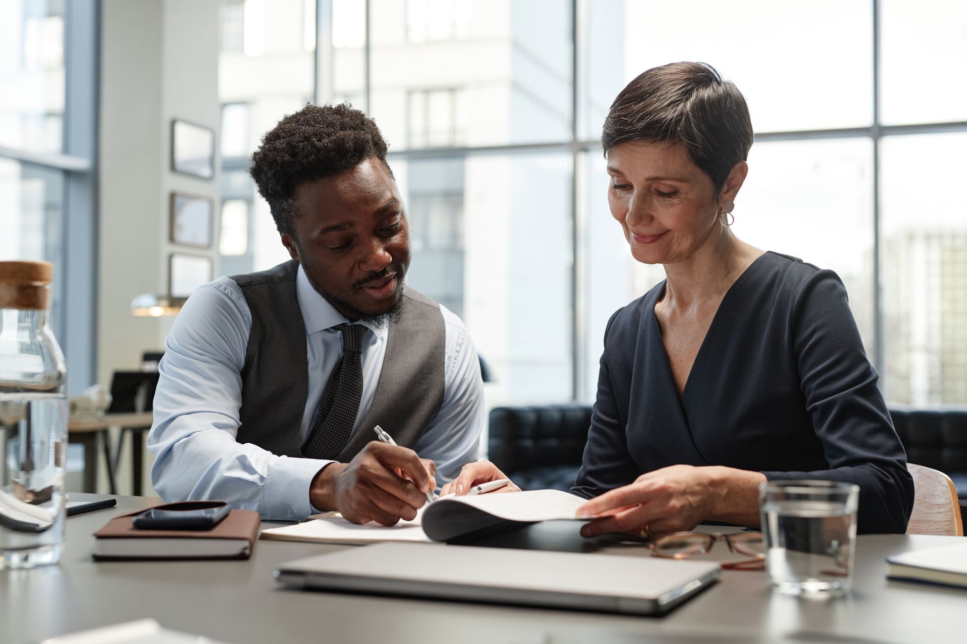 Man in vest writing, woman in blue dress looking at papers. Office setting, natural light.