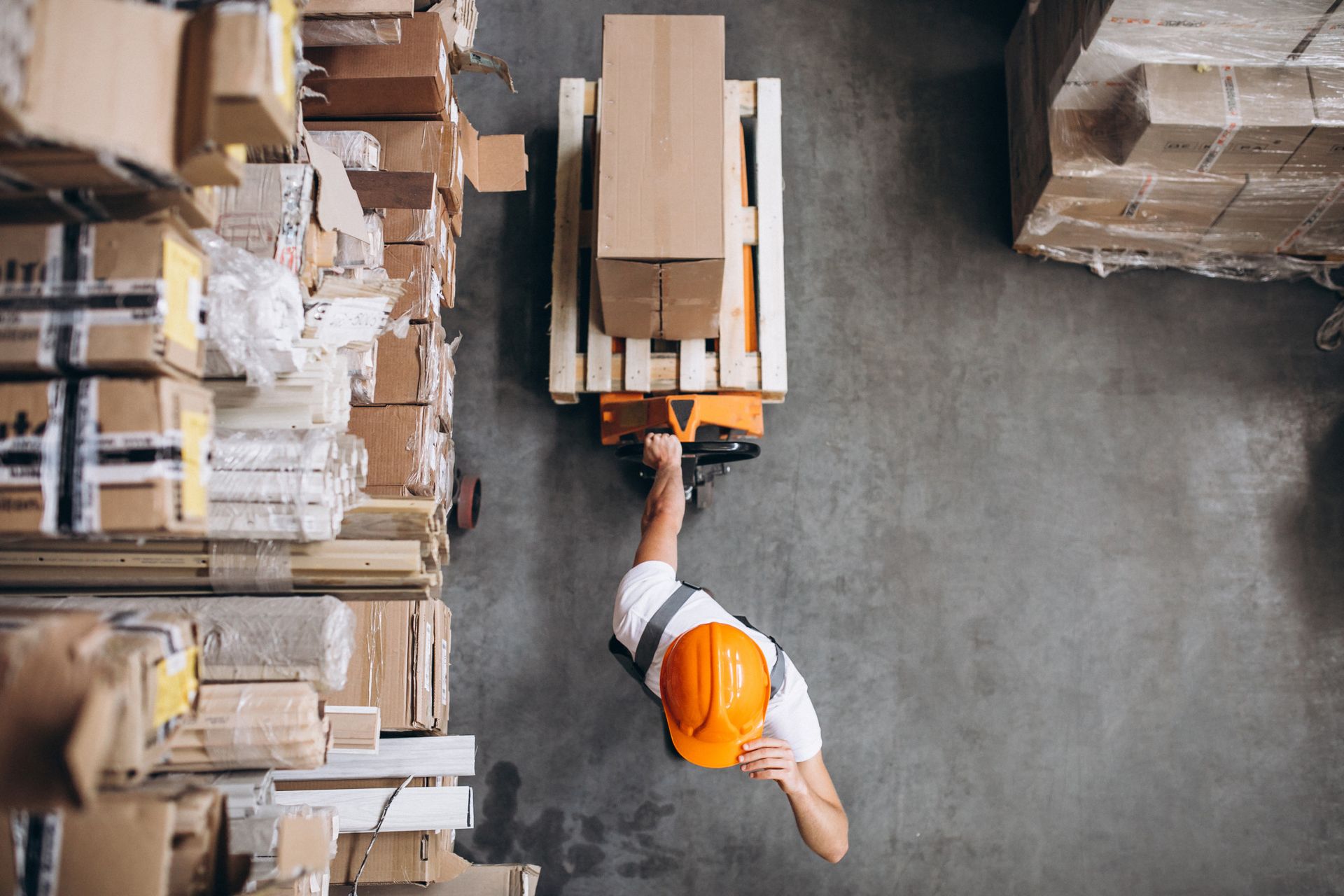 Warehouse worker in orange hard hat operating a pallet jack, moving a cardboard box, from an overhead perspective.