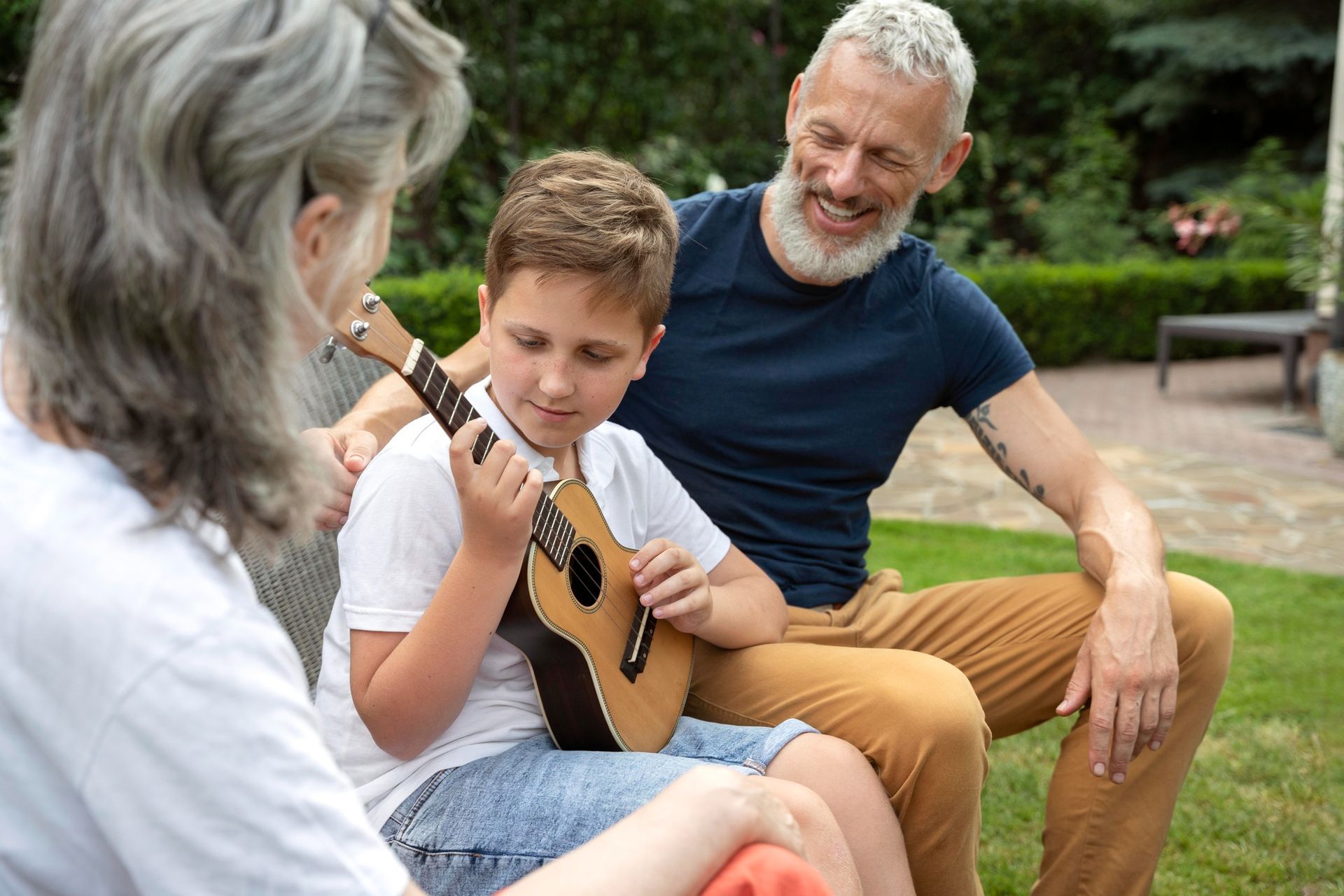 Boy playing ukulele for grandparents in outdoor setting.