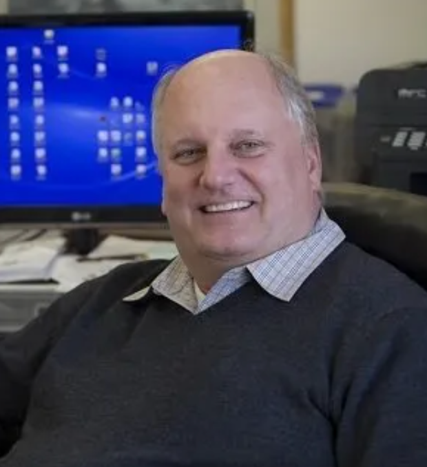 Man with a bald head, smiling, seated in front of a computer. Wearing a gray sweater, light-colored shirt collar visible.