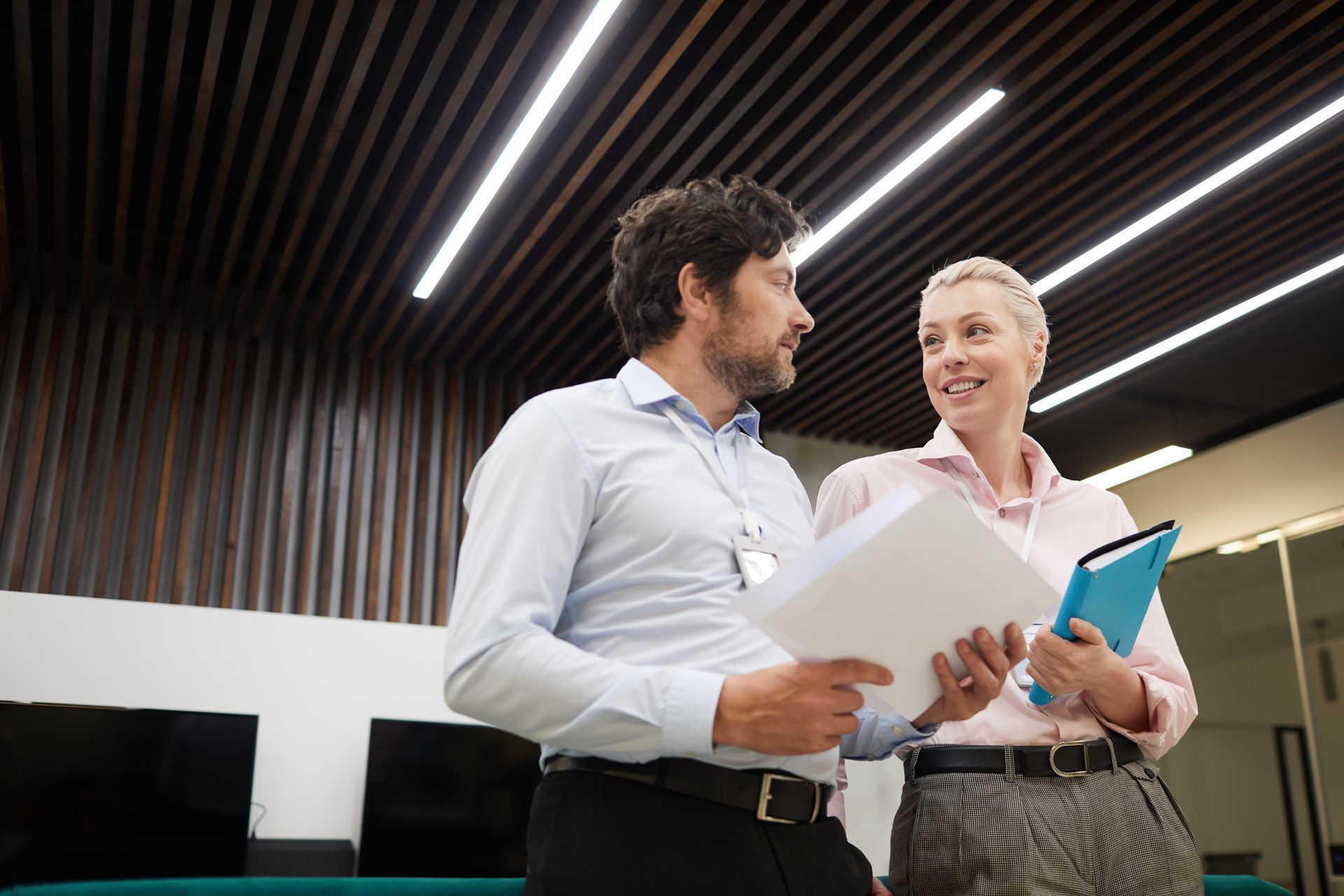 Two people in business attire, looking at documents, in an office with a wood ceiling and overhead lights.