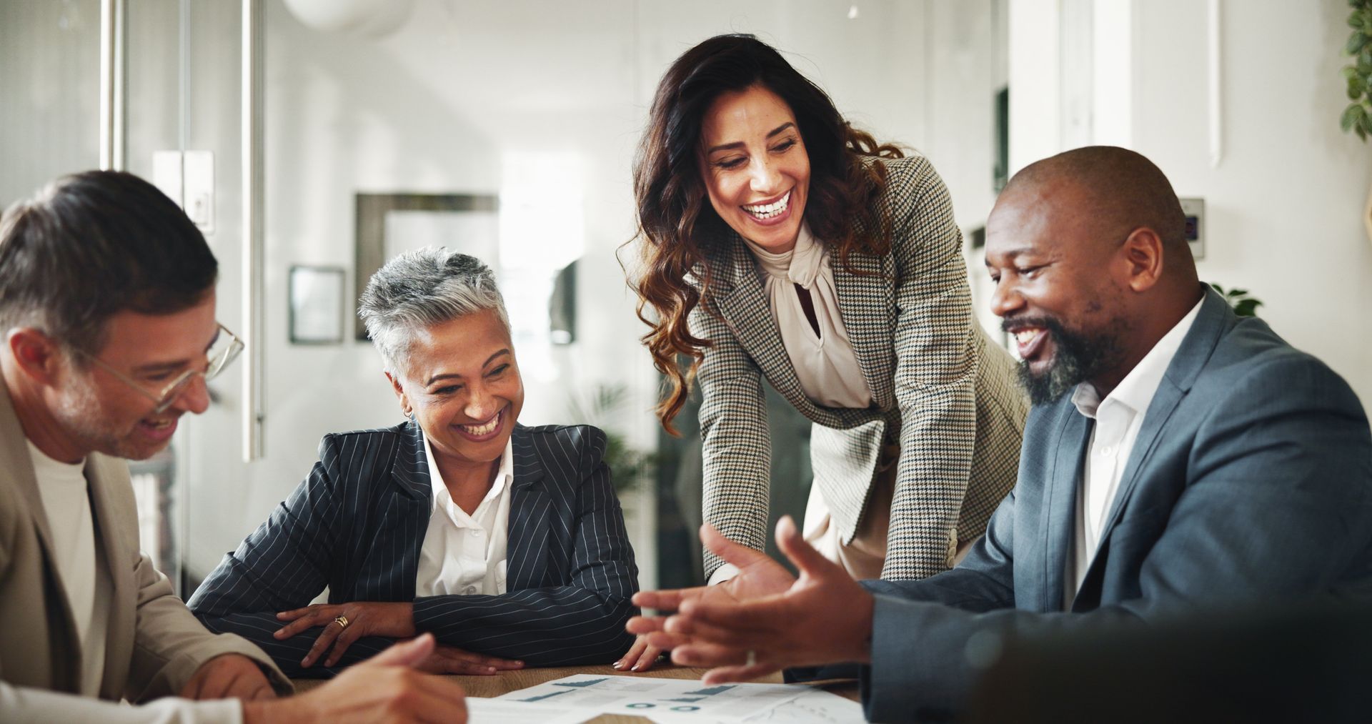 Group of people smiling, collaborating around a table in a modern office, discussing documents.