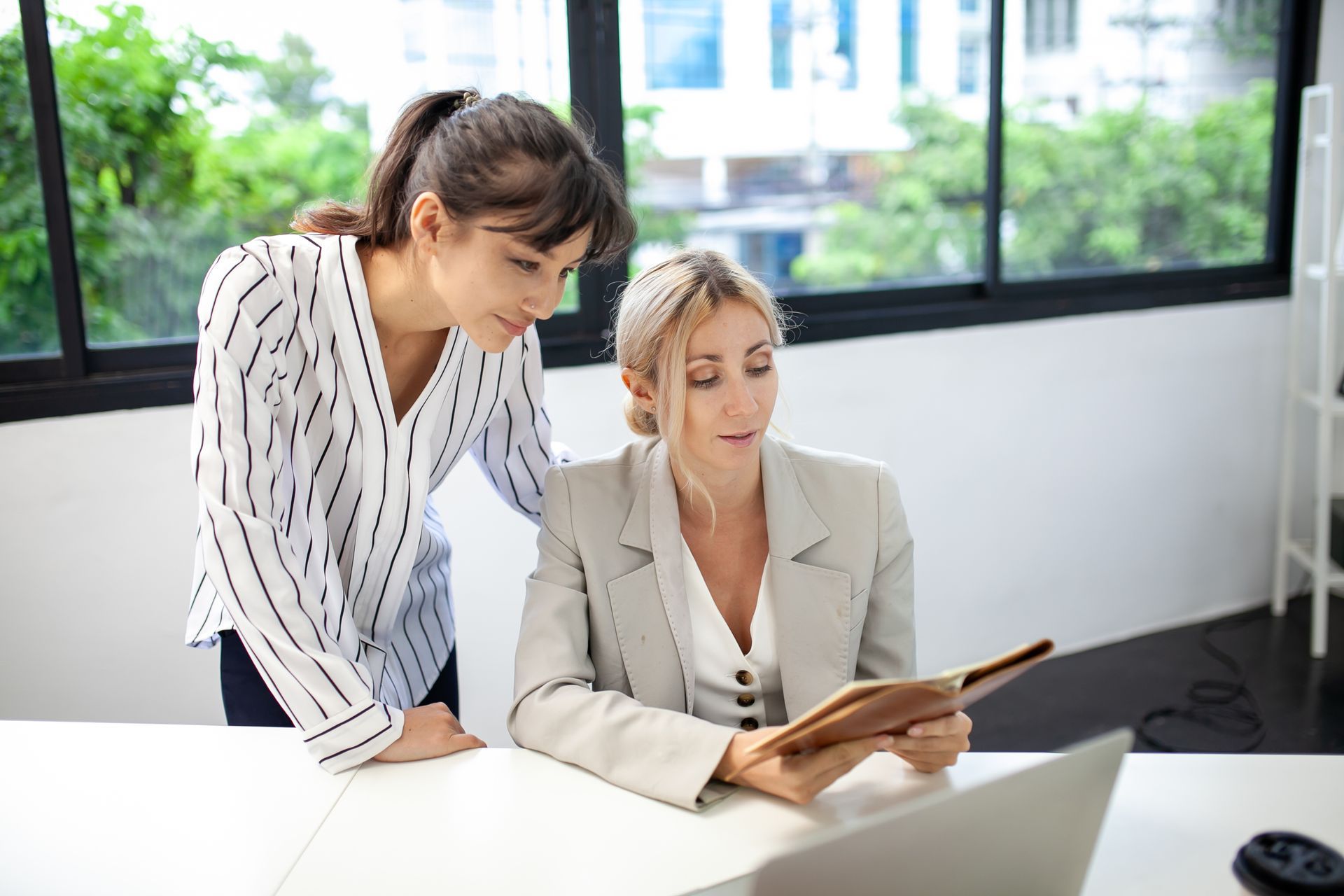 Two women in an office, one looking over the shoulder of the other, who is looking at a notebook.