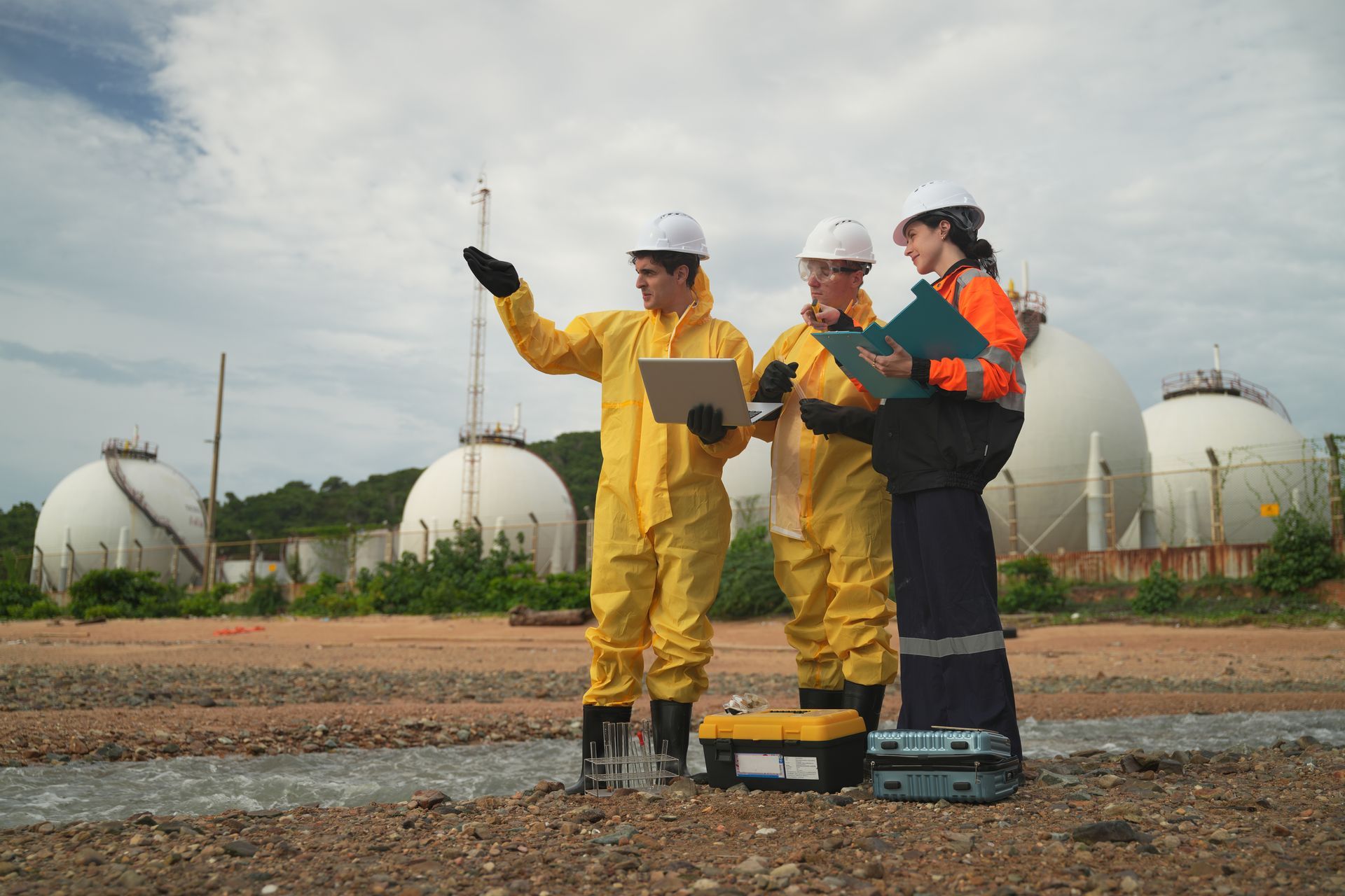 Three environmental workers in hazmat suits assessing a site near storage tanks.