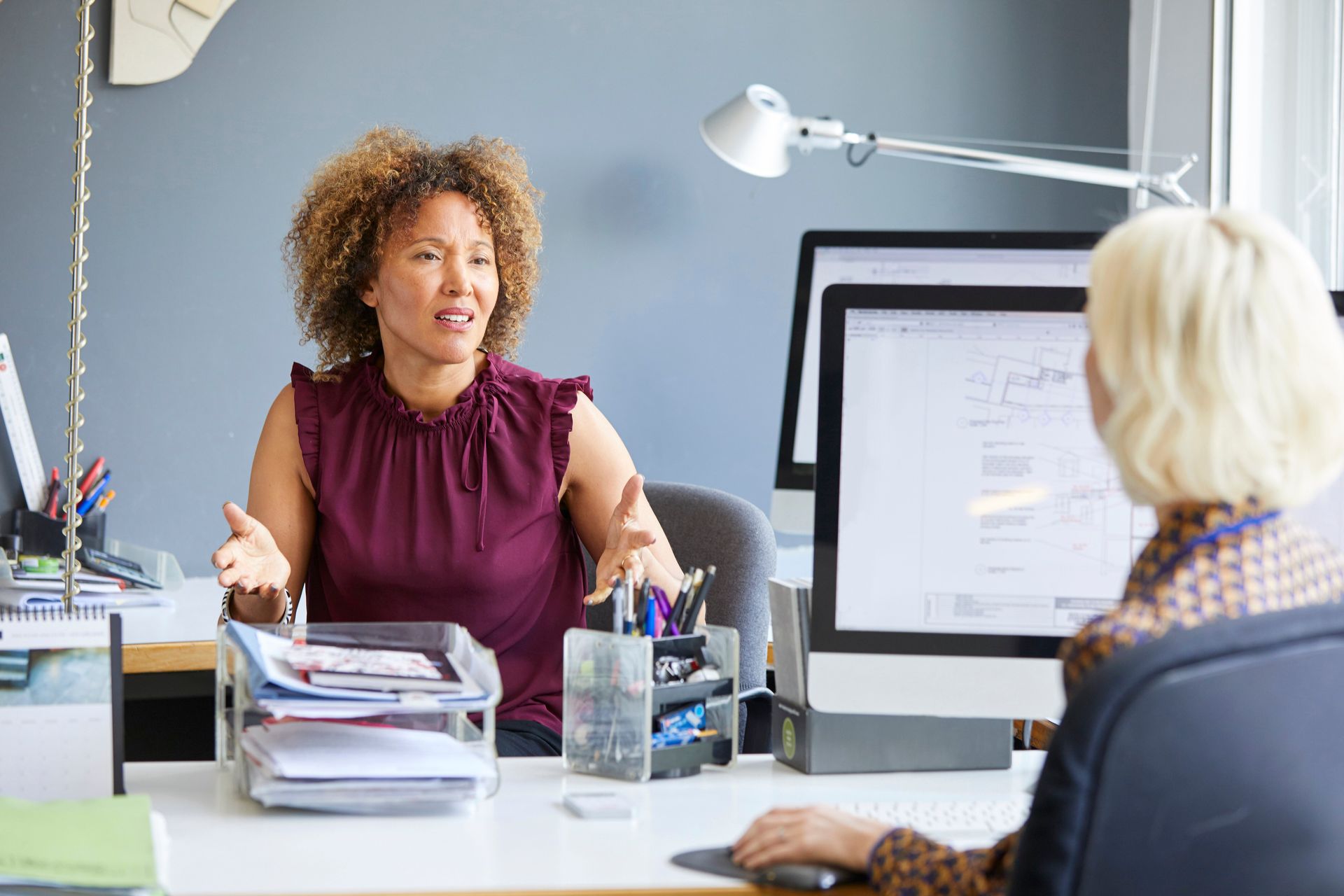 Woman in maroon top talking to another woman in an office setting with computer screens and desk supplies.