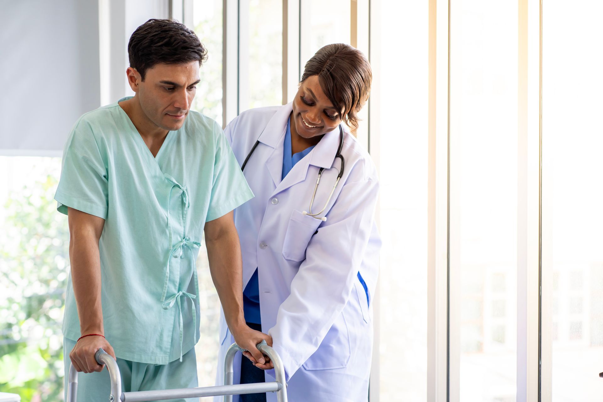 Man in hospital gown using a walker, assisted by a smiling doctor indoors.