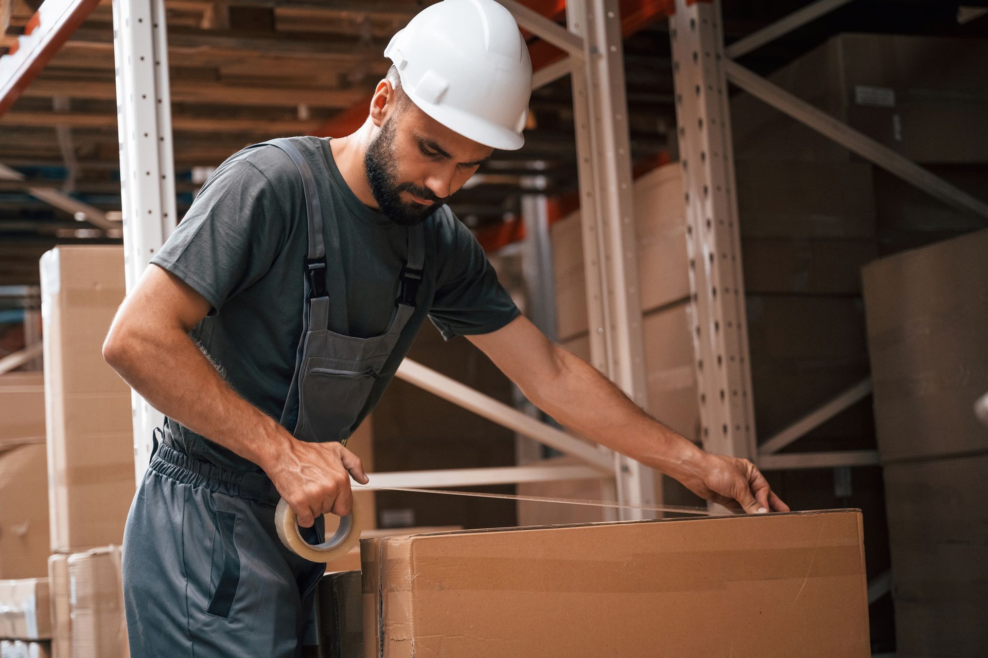 Warehouse worker in a white hard hat taping a cardboard box on a shelf.