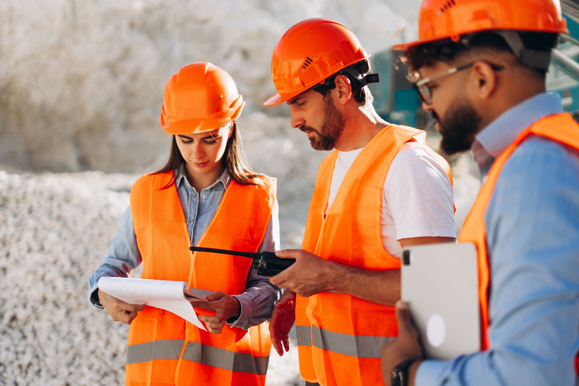 Construction workers in orange safety vests and helmets review blueprints and digital tablet on site.