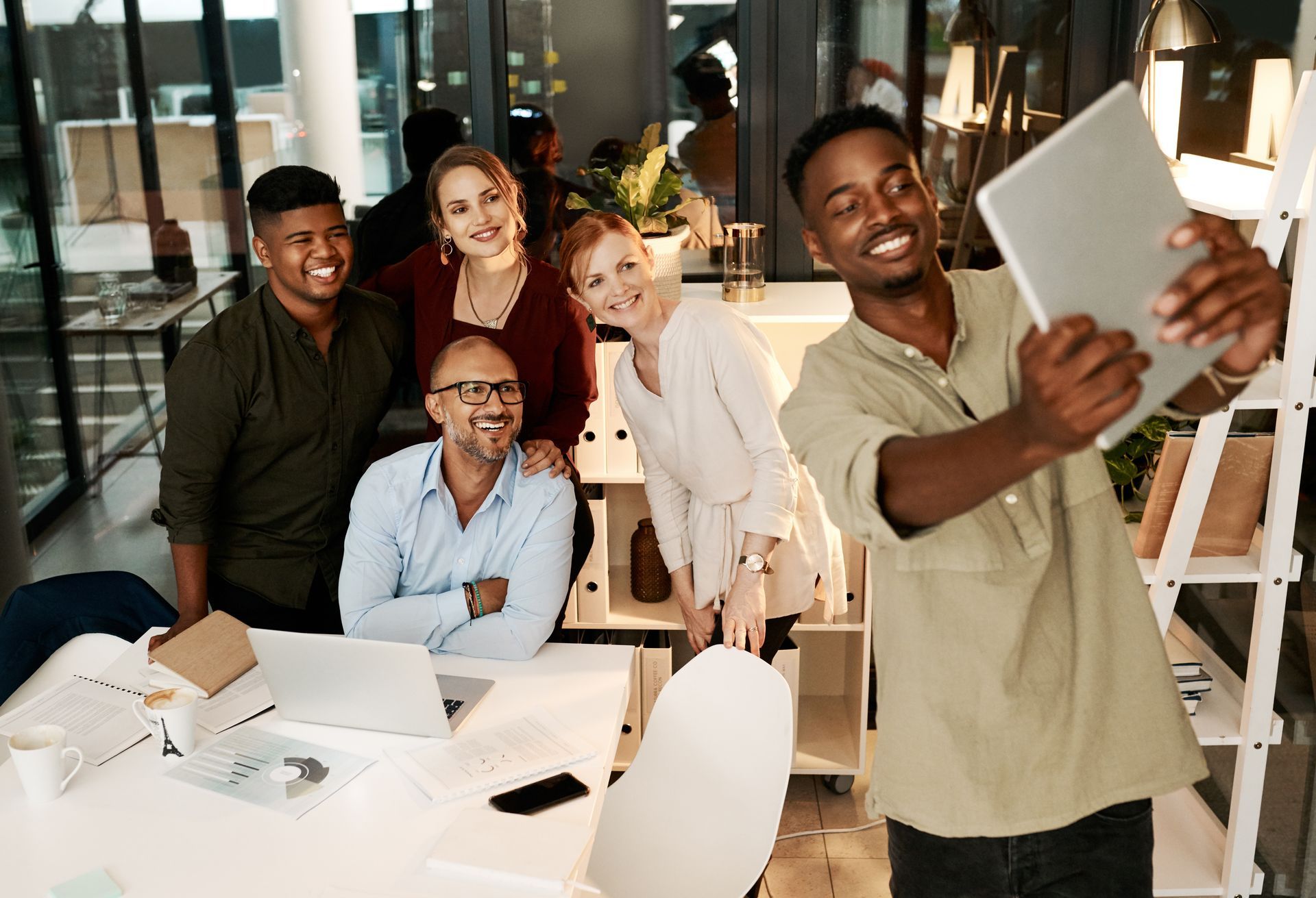 A group of people in an office smiling, one taking a selfie with a tablet.