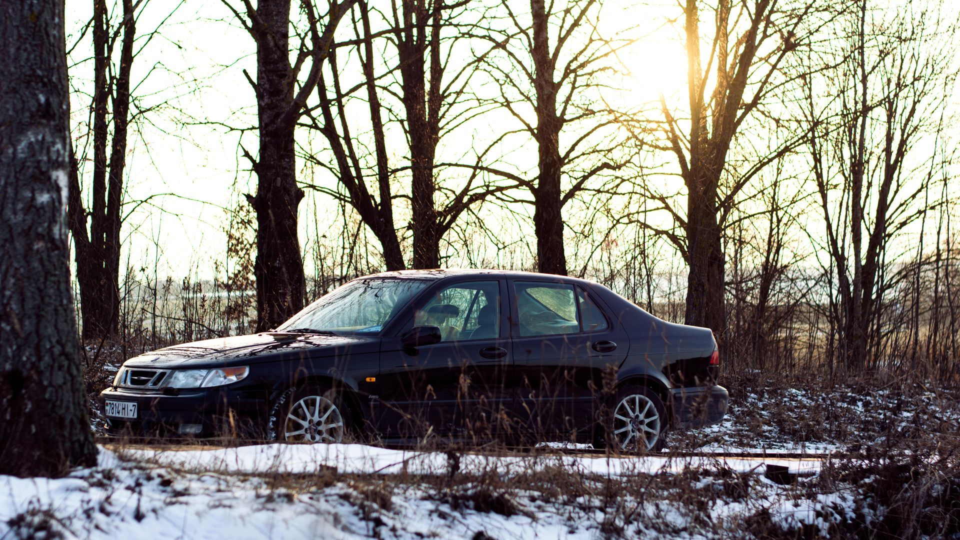 Black Saab sedan parked on snowy roadside with trees in background.