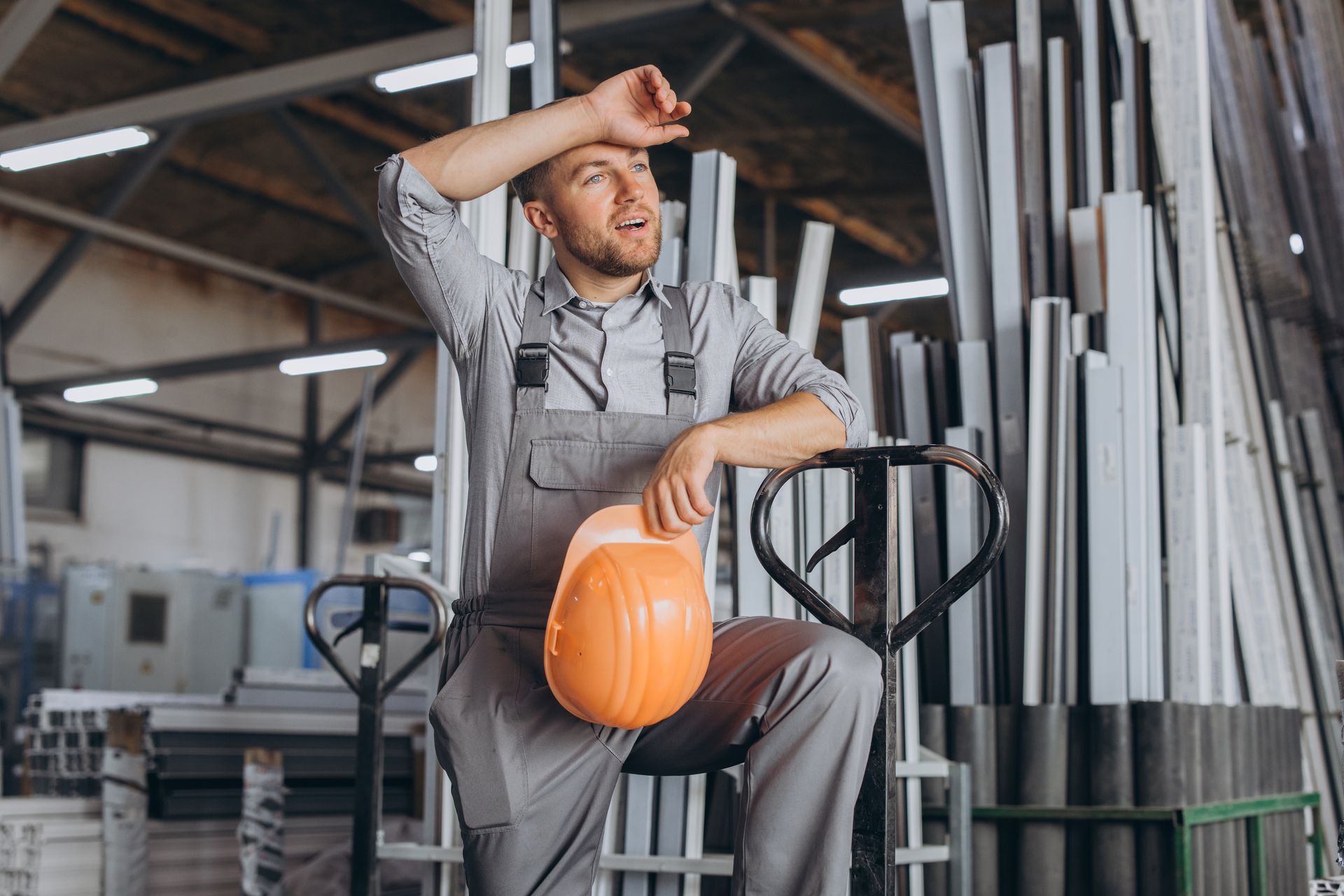 Man in gray overalls wipes sweat, holding an orange hard hat, leaning against a forklift in a warehouse.